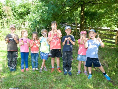 Eight children stand outdoors on grass in a row, smiling and holding up sticks. They are dressed casually, some wearing hats, and there are trees and a wooden fence in the background.