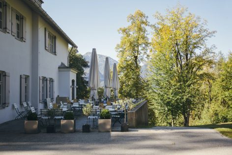Outdoor terrace of a building with white tables and chairs, large closed umbrellas, and potted plants, surrounded by green trees and mountains visible in the background under a clear sky.