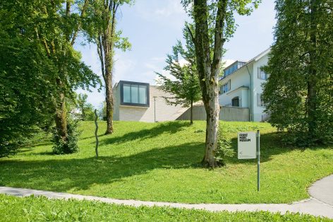 A modern building stands behind trees on a grassy hill, with a sign in the foreground reading Franz Marc Museum and an arrow pointing left. The sky is clear and sunny.