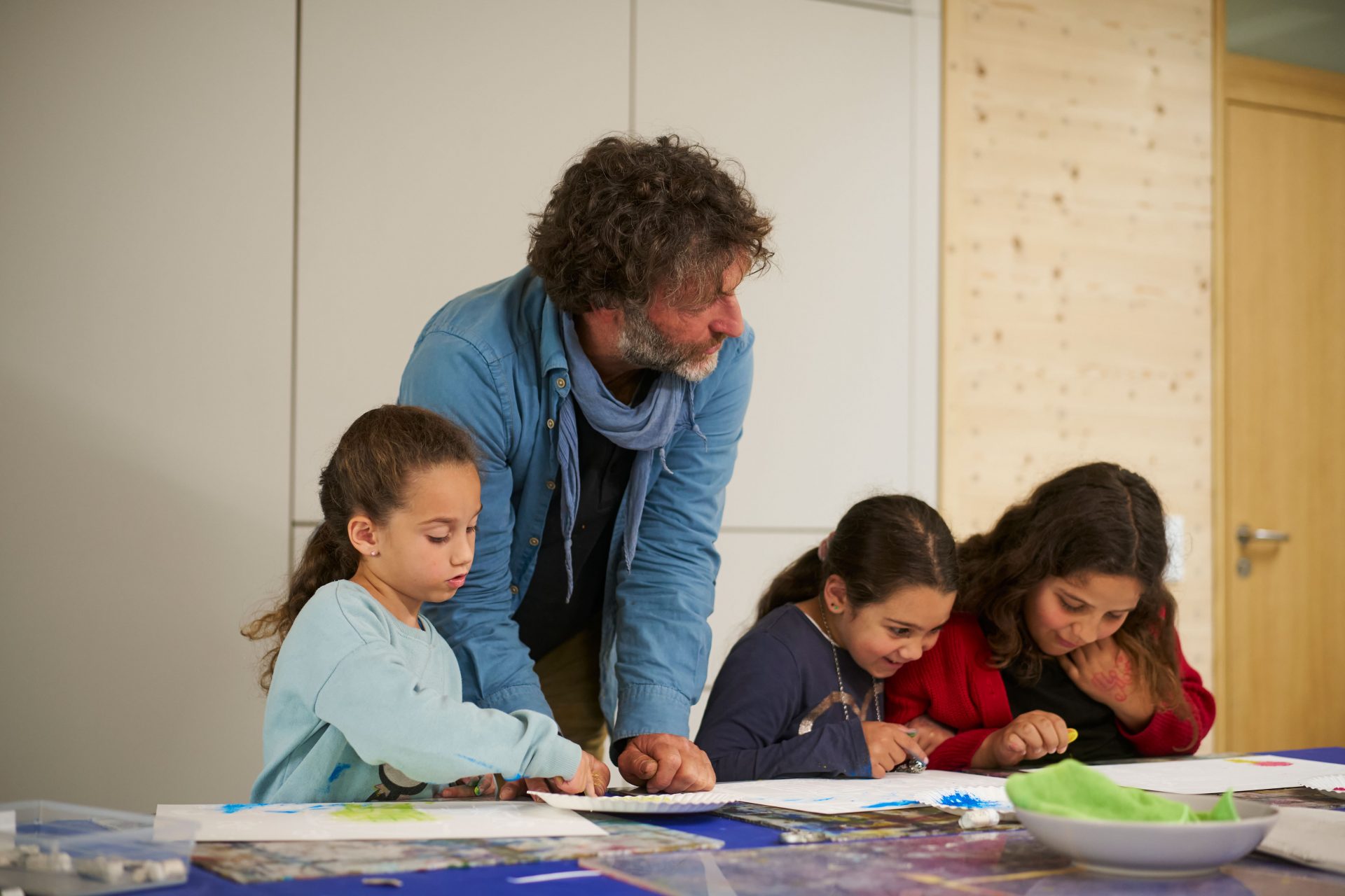 Ein Mann beugt sich über einen Tisch und hilft drei jungen Mädchen bei einem Kunstprojekt, während sie in einem hellen, modernen Klassenzimmer auf Papier zeichnen und malen.
