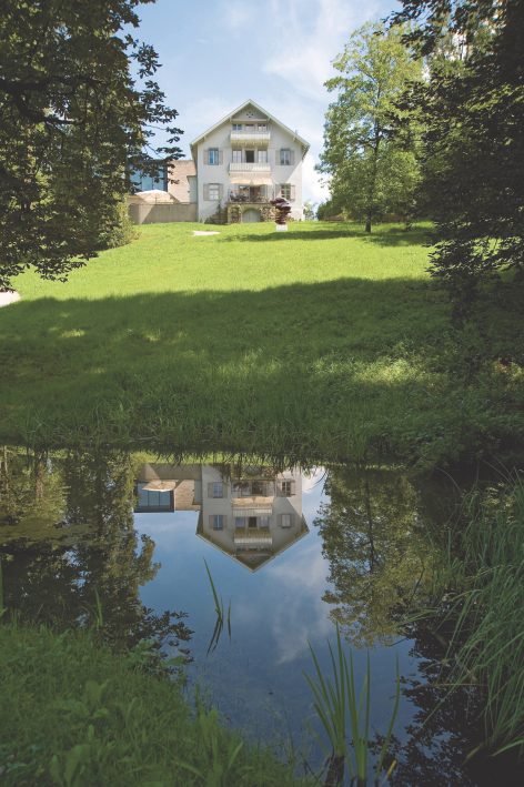 A white house with balconies sits atop a grassy hill, reflected clearly in a small, calm pond surrounded by green grass and trees under a blue sky.