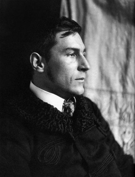 Black and white portrait of a man in profile, wearing a dark coat with a textured collar, a white shirt, and a patterned tie, looking to the right with a neutral expression.