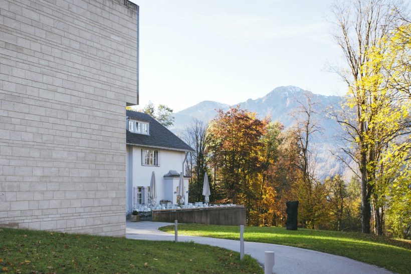 Ein modernes Gebäude mit hellen Ziegelwänden steht neben einem weißen Haus mit Sitzgelegenheiten im Freien, umgeben von herbstlichen Bäumen und Bergen im Hintergrund unter einem klaren Himmel.