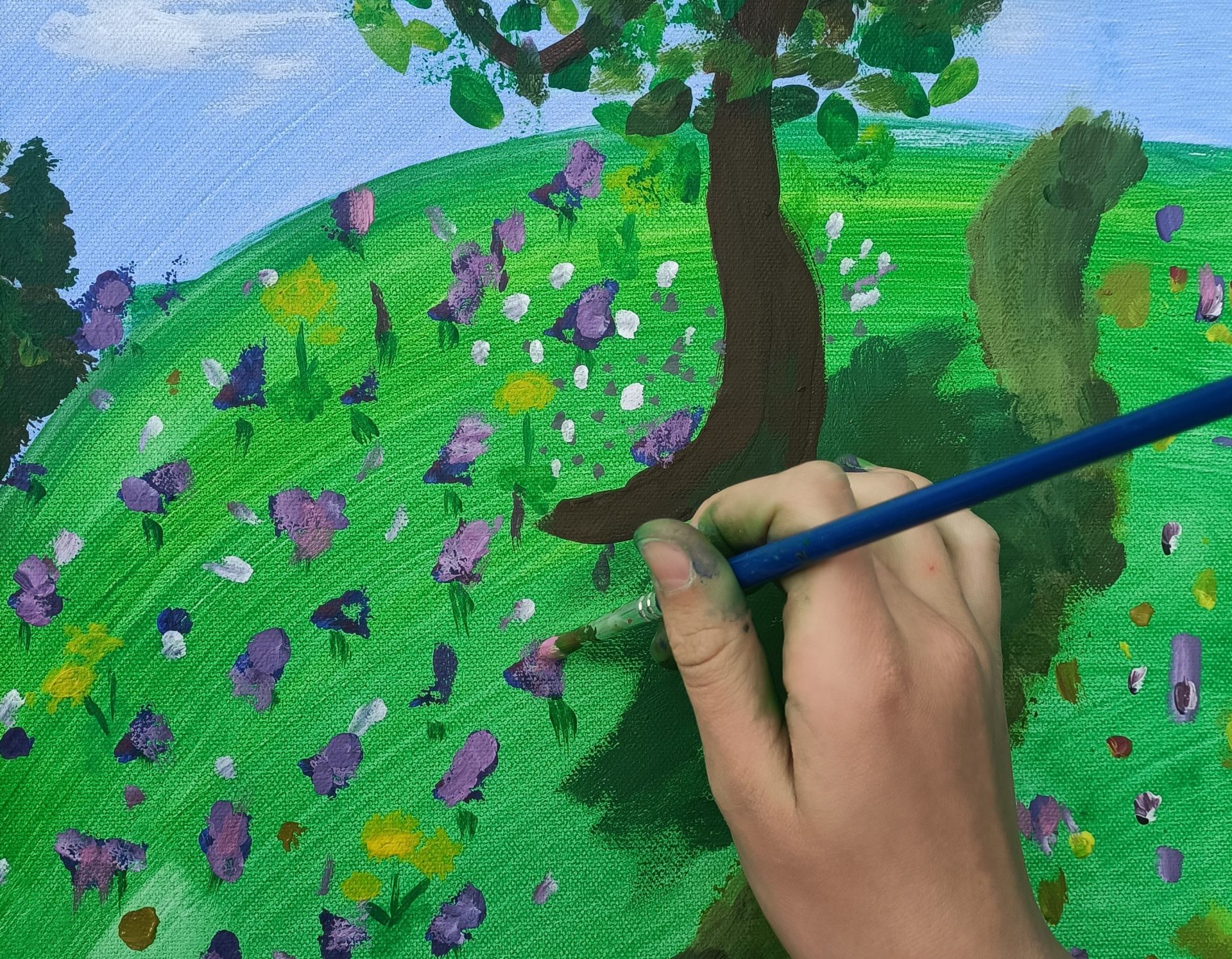 Eine Hand hält einen Pinsel und malt eine bunte Landschaft mit einem Baum und Blumen auf grünem Gras unter einem blauen Himmel auf eine Leinwand.