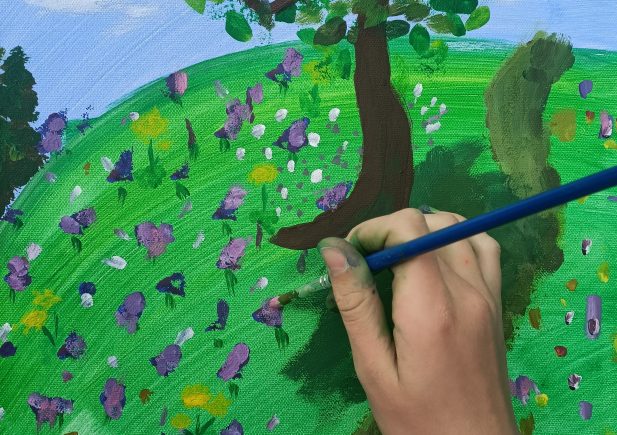 Eine Hand hält einen Pinsel und malt eine bunte Landschaft mit einem Baum und Blumen auf grünem Gras unter einem blauen Himmel auf eine Leinwand.