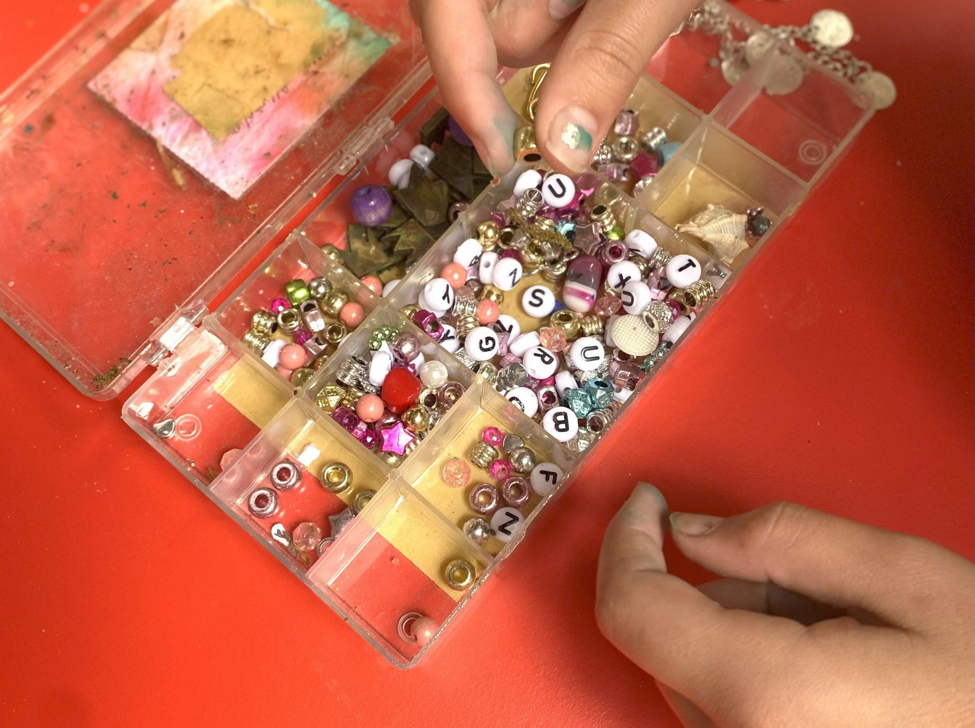 Eine Person wählt Buchstabenperlen aus einem offenen Kunststoffbehälter aus, der mit bunten Perlen und Zubehör für die Schmuckherstellung auf einer roten Fläche gefüllt ist.