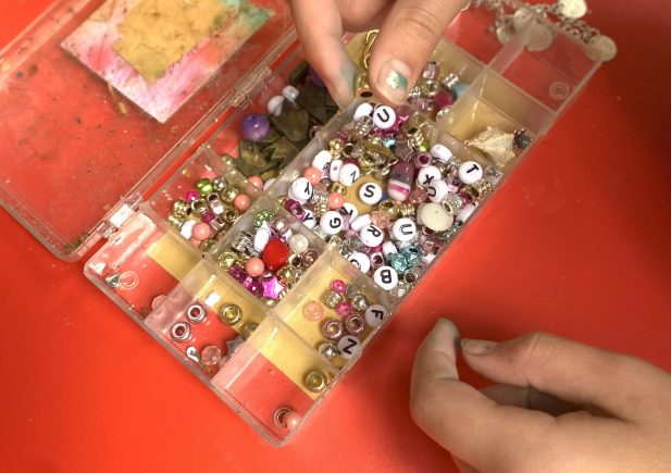 Eine Person wählt Buchstabenperlen aus einem offenen Kunststoffbehälter aus, der mit bunten Perlen und Zubehör für die Schmuckherstellung auf einer roten Fläche gefüllt ist.