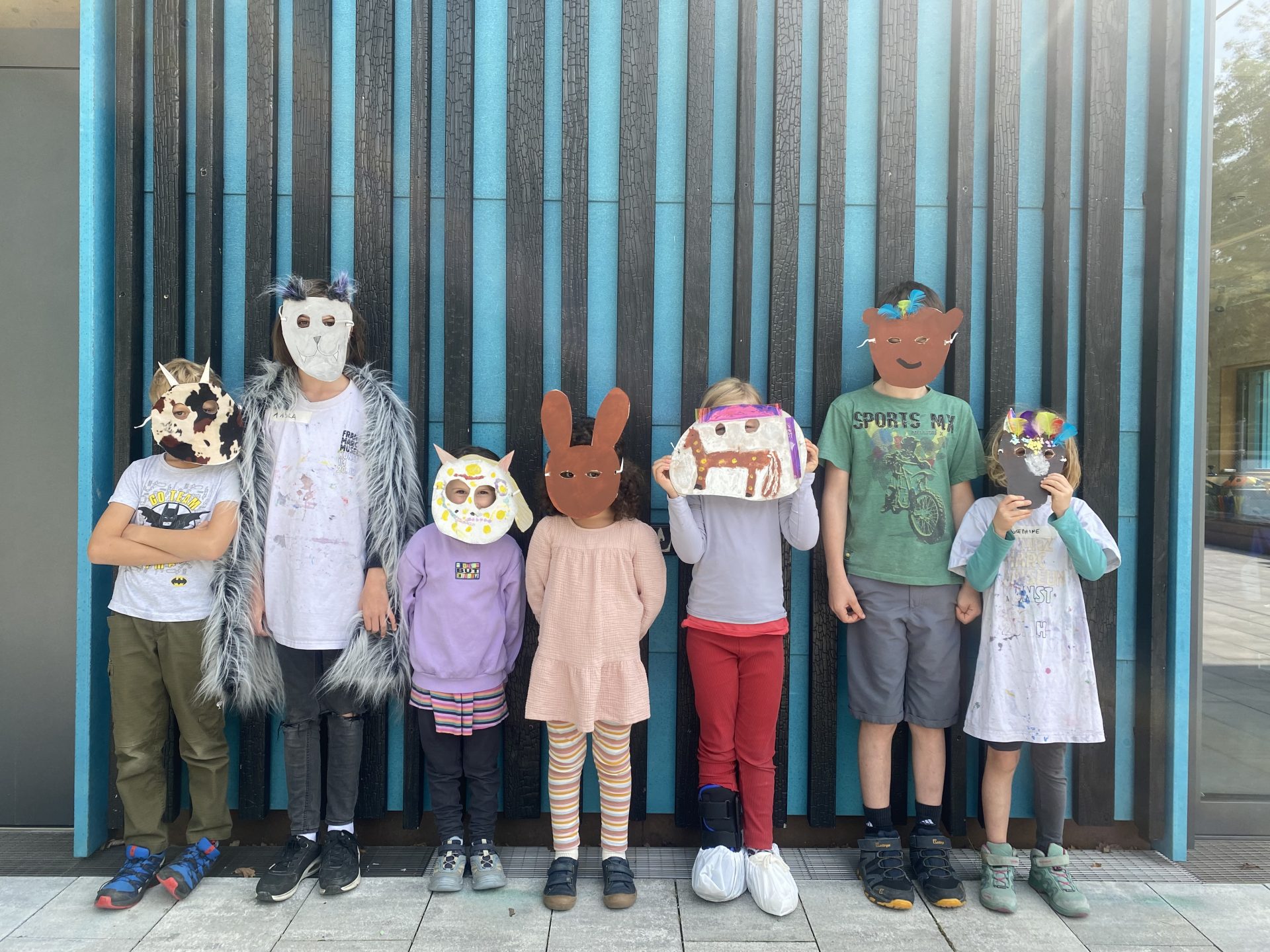 Sieben Kinder stehen in einer Reihe und tragen jeweils eine handgefertigte Tier- oder Figurenmaske. Sie stehen im Freien vor einer blau gestreiften Wand, sind leger gekleidet und ihre Gesichter sind vollständig von den Masken verdeckt.