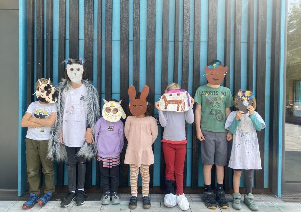 Sieben Kinder stehen in einer Reihe und tragen jeweils eine handgefertigte Tier- oder Figurenmaske. Sie stehen im Freien vor einer blau gestreiften Wand, sind leger gekleidet und ihre Gesichter sind vollständig von den Masken verdeckt.