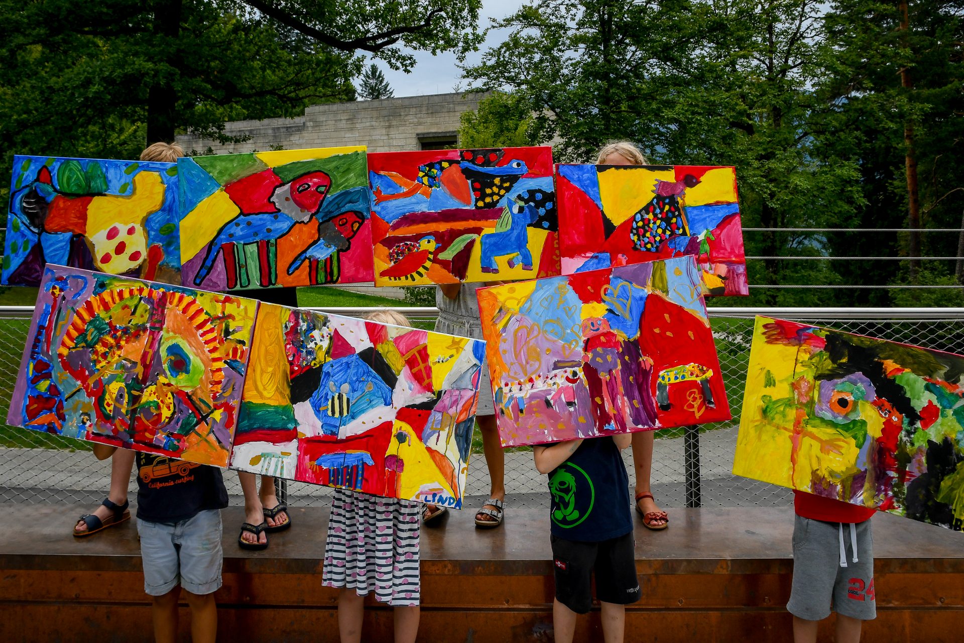 Fünf Kinder stehen im Freien und halten bunte, abstrakte Gemälde hoch, die ihre Gesichter bedecken. Die lebendigen Kunstwerke zeigen kühne Formen, Tiere und Muster. Im Hintergrund sind Bäume und ein modernes Gebäude zu sehen.