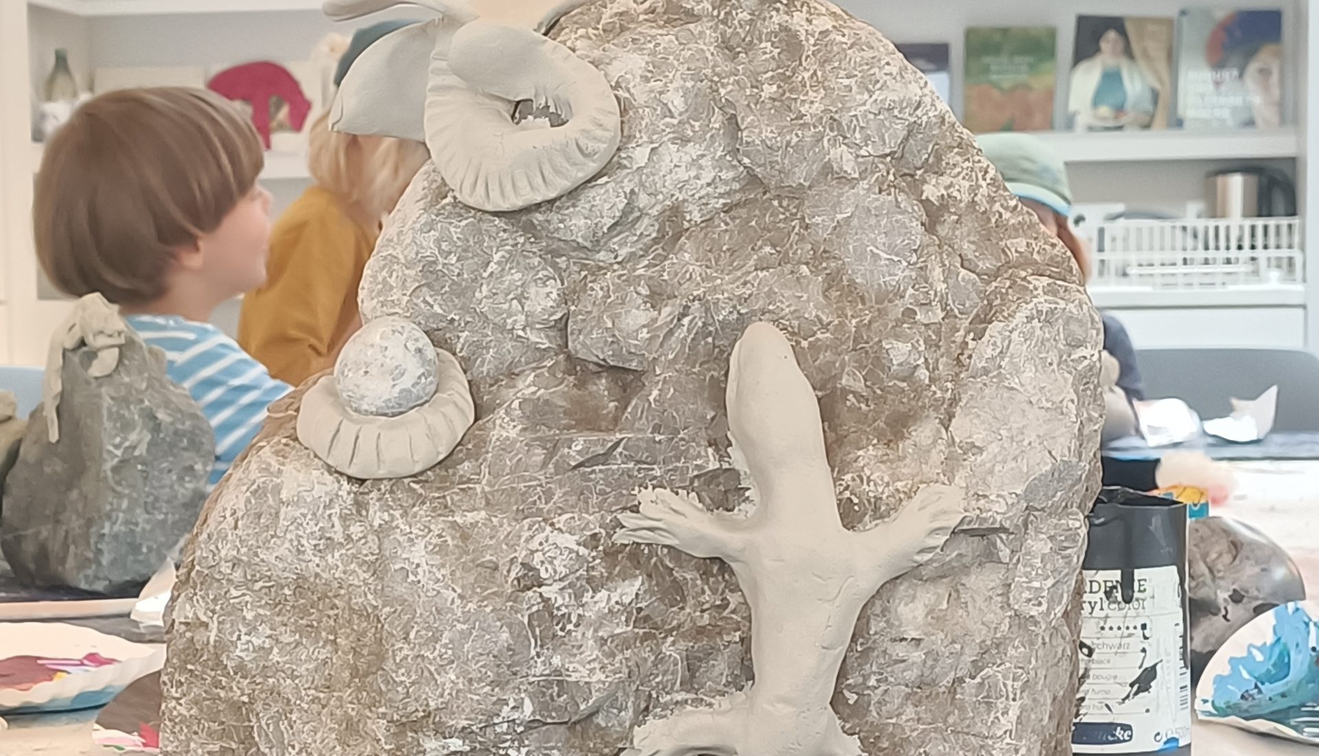 Auf einem großen Felsen stehen Tonfiguren einer Eidechse und zweier Ammoniten. Im Hintergrund sitzen Kinder an einem Tisch, hinter dem in Regalen Kunstsachen und Bilder zu sehen sind.