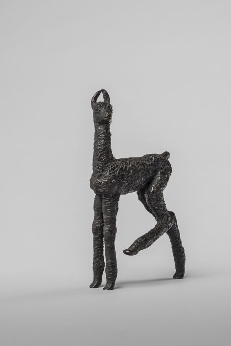 A small, textured bronze sculpture of a standing llama with elongated legs and neck, shown against a plain light gray background.