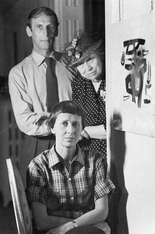 Drei Erwachsene posieren in einem Innenraum: Ein Mann mit Hemd und Krawatte steht hinter zwei Frauen, von denen die eine einen geblümten Hut und ein gepunktetes Kleid trägt, während die andere in einem karierten Hemd davor sitzt. An der Wand neben ihnen hängt ein abstraktes Gemälde.