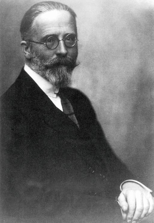 Schwarz-Weiß-Porträt des Sammlers Rudolf Ibachs mit Brille, Vollbart und Schnurrbart, der Anzug und Krawatte trägt, sitzend und leicht zur Seite blickend vor einem schlichten Hintergrund.