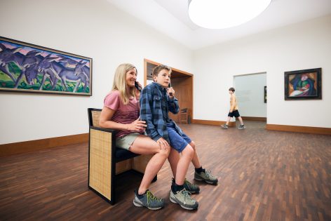 A woman and a boy sit together on a bench in an art gallery, looking at artwork on the walls, while another boy walks in the background. The room has wood flooring and modern paintings displayed.
