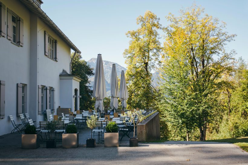 Eine Restaurantterrasse im Freien mit leeren Tischen und Stühlen neben einem weißen Gebäude, umgeben von üppigen Bäumen und Bergen in der Ferne unter einem klaren blauen Himmel.