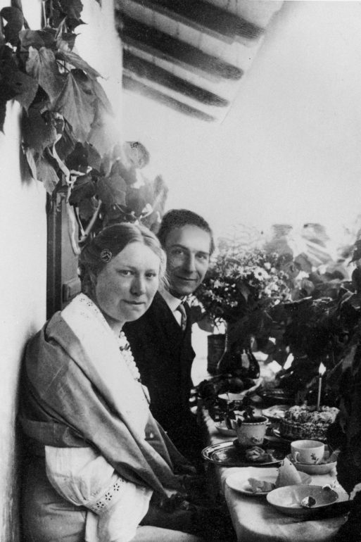 Schwarz-Weiß-Foto einer Frau und eines Mannes, die zusammen an einem kleinen Tisch im Freien mit Teetassen sitzen, umgeben von Pflanzen und Blumen. Die Frau schaut in die Kamera, während der Mann lächelt. Beide tragen Kleidung aus dem frühen 20.