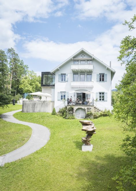 A white, three-story house with balconies sits on a green lawn under a blue sky. A curved path leads to the house, and a modern, abstract sculpture stands in the yard. Trees surround the property.