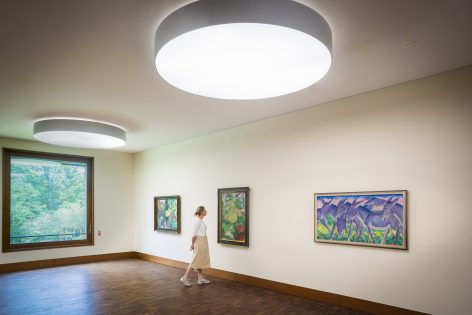 A person walks through a modern art gallery with large circular ceiling lights, wooden floors, and several colorful paintings on cream-colored walls. A large window shows greenery outside.