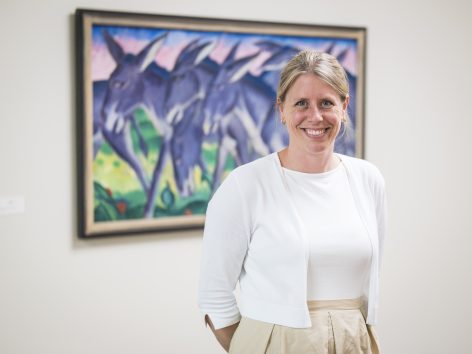 A woman with blonde hair tied back, wearing a white top and beige skirt, stands smiling in front of a colorful painting of blue donkeys on a wall.