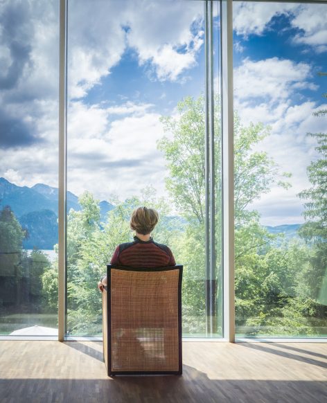 A person sits in a chair facing large floor-to-ceiling windows, looking out at green trees, mountains, and a partly cloudy sky. Natural light fills the modern, serene space.