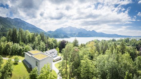 A modern building with a green roof sits among dense trees, overlooking a lake with mountains in the background under a partly cloudy sky.