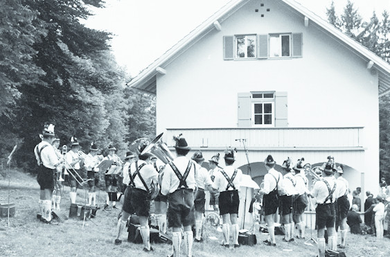 Eine Gruppe von Musikern in traditioneller bayerischer Kleidung spielt auf einem Schwarz-Weiß-Foto vor einem weißen Haus mit Fensterläden, das von Bäumen umgeben ist, Instrumente.