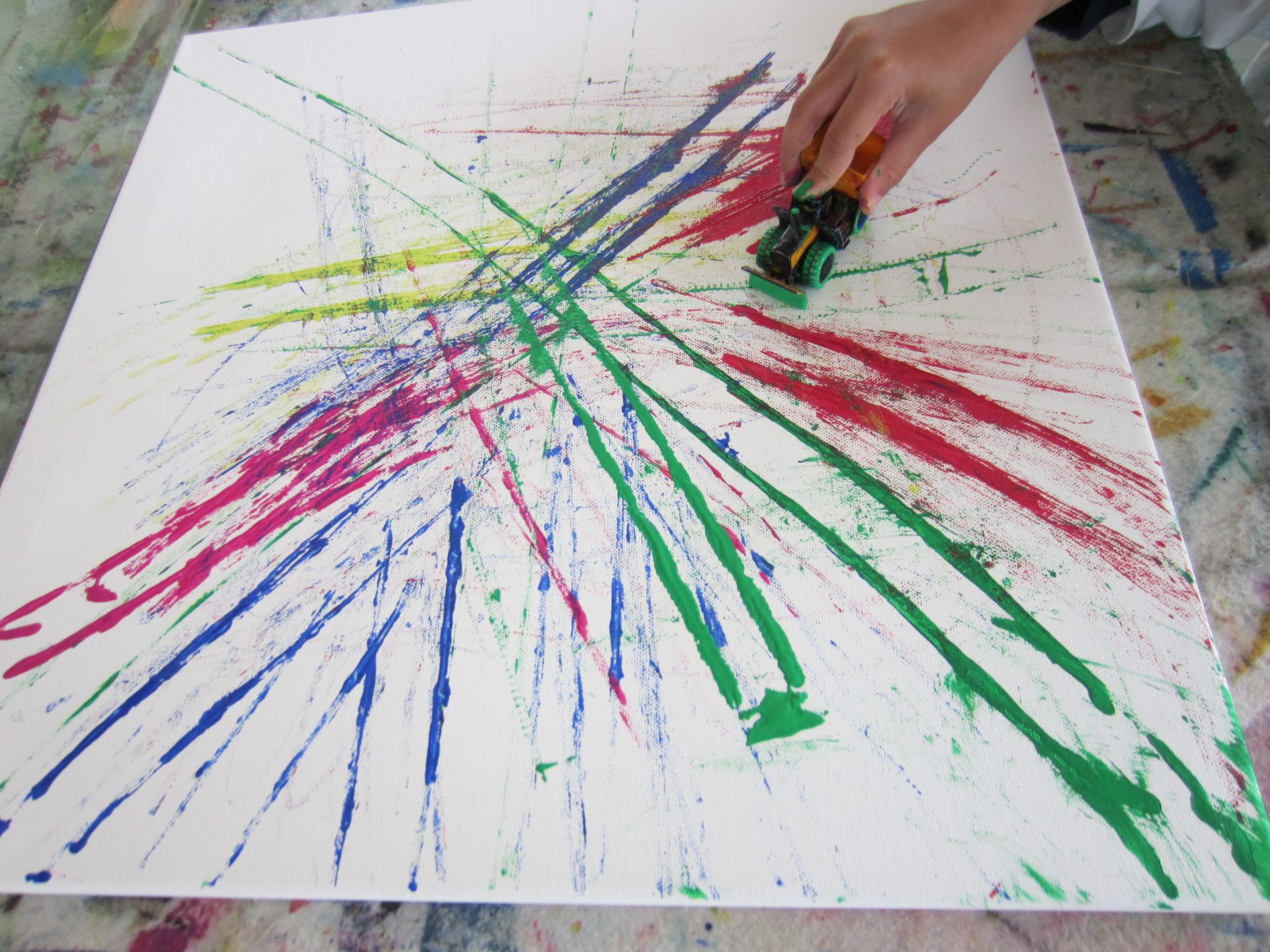 Eine Hand benutzt ein Spielzeugauto, um leuchtend rote, grüne, gelbe und blaue Farbe auf einer weißen Leinwand zu verteilen, wodurch bunte, sich kreuzende Schlieren und Spritzer entstehen.
