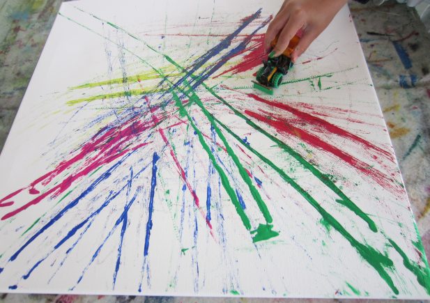Eine Hand benutzt ein Spielzeugauto, um leuchtend rote, grüne, gelbe und blaue Farbe auf einer weißen Leinwand zu verteilen, wodurch bunte, sich kreuzende Schlieren und Spritzer entstehen.