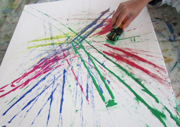 Eine Hand benutzt ein Spielzeugauto, um leuchtend rote, grüne, gelbe und blaue Farbe auf einer weißen Leinwand zu verteilen, wodurch bunte, sich kreuzende Schlieren und Spritzer entstehen.