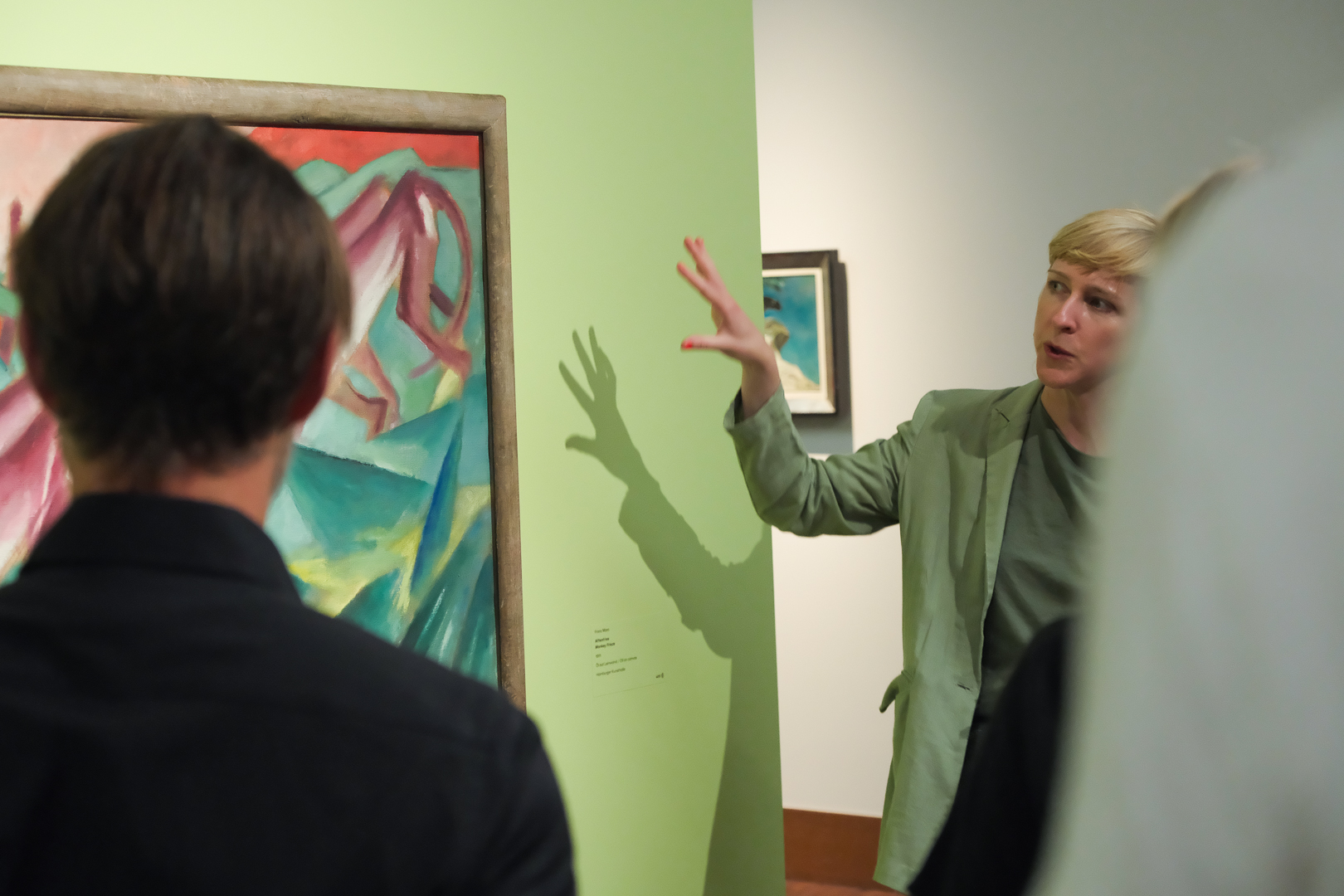 Eine Person in einem hellgrünen Anzug gestikuliert, während sie zu einer kleinen Gruppe vor einem abstrakten Gemälde spricht, das auf einer hellgrünen Wand in einer Kunstgalerie ausgestellt ist.