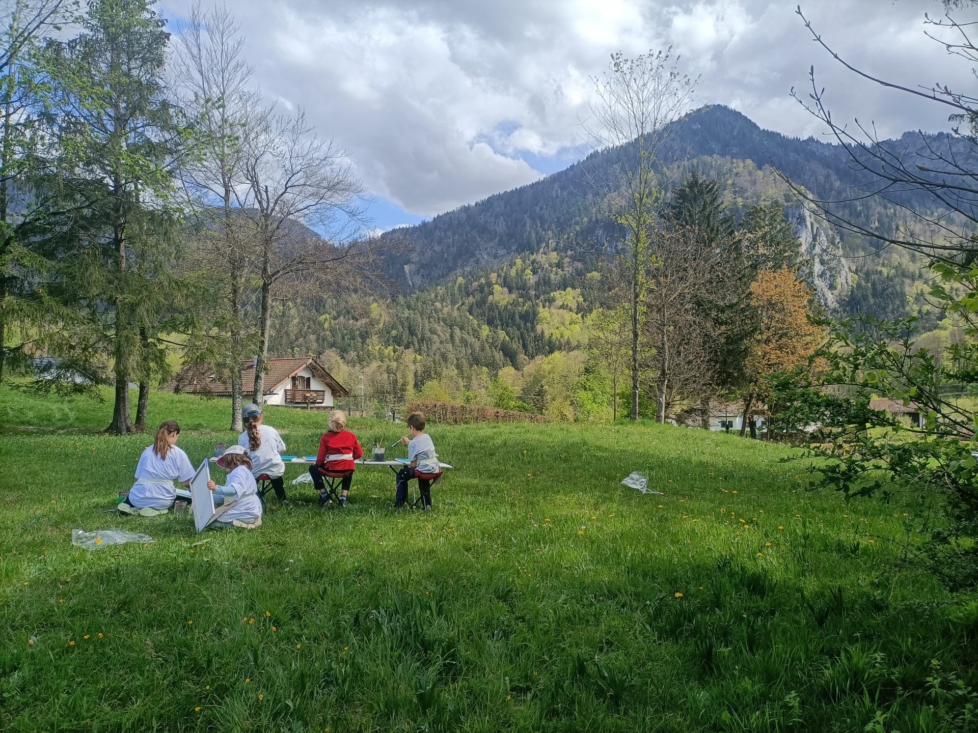 Eine Gruppe von Menschen sitzt auf Stühlen und Decken und zeichnet oder malt im Freien auf einer grasbewachsenen Wiese mit Bergen, Bäumen und Häusern im Hintergrund unter einem teilweise bewölkten Himmel.