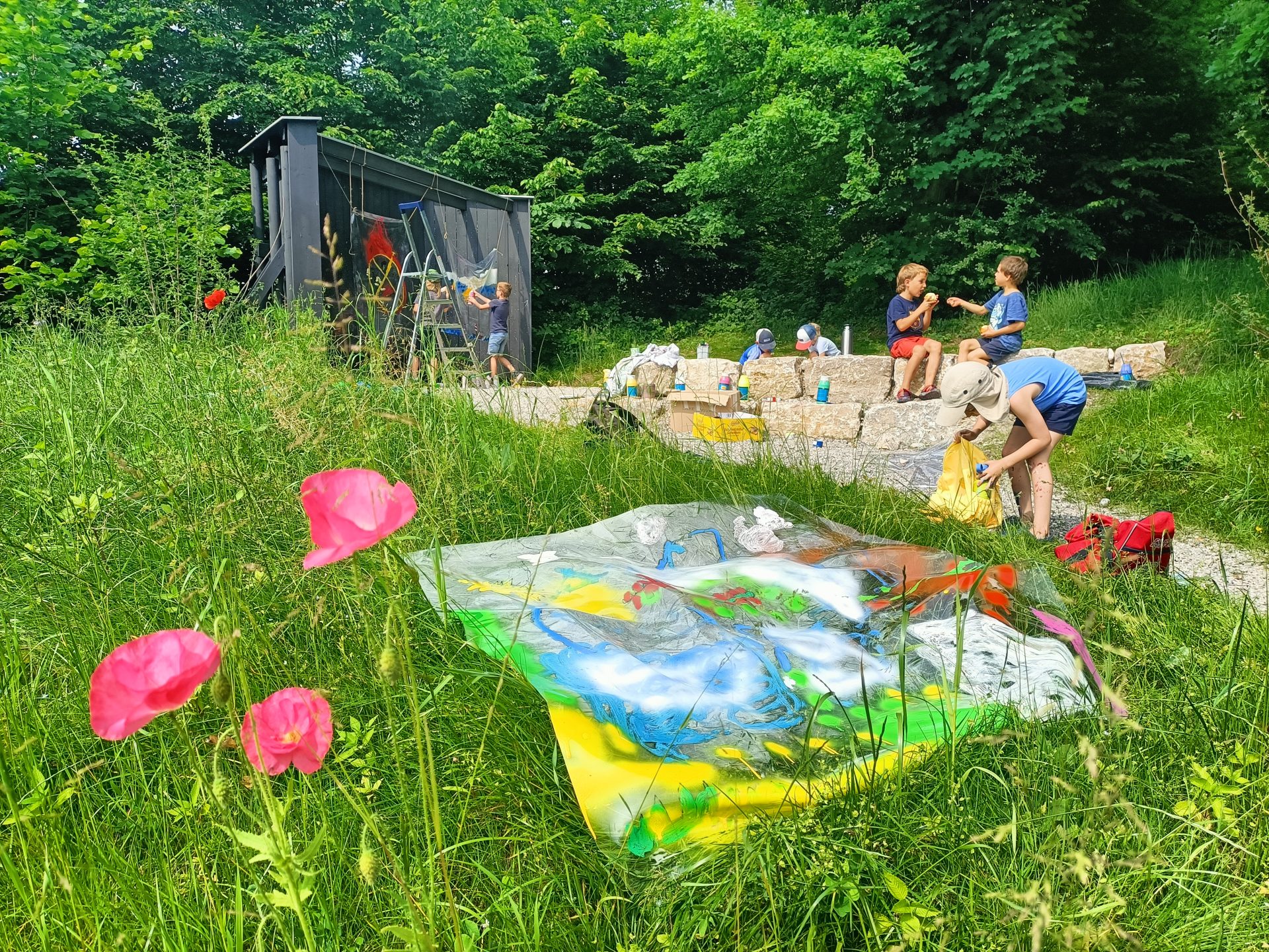 Kinder malen im Freien auf Steinstufen, umgeben von grünem Gras und Bäumen. Helle Kunstwerke auf transparentem Plastik liegen auf dem Gras, mit rosa Blumen im Vordergrund und einem kleinen Unterstand im Hintergrund.