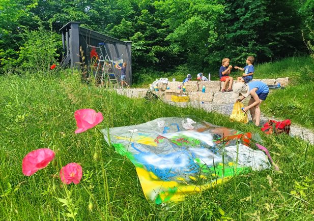 Kinder malen im Freien auf Steinstufen, umgeben von grünem Gras und Bäumen. Helle Kunstwerke auf transparentem Plastik liegen auf dem Gras, mit rosa Blumen im Vordergrund und einem kleinen Unterstand im Hintergrund.