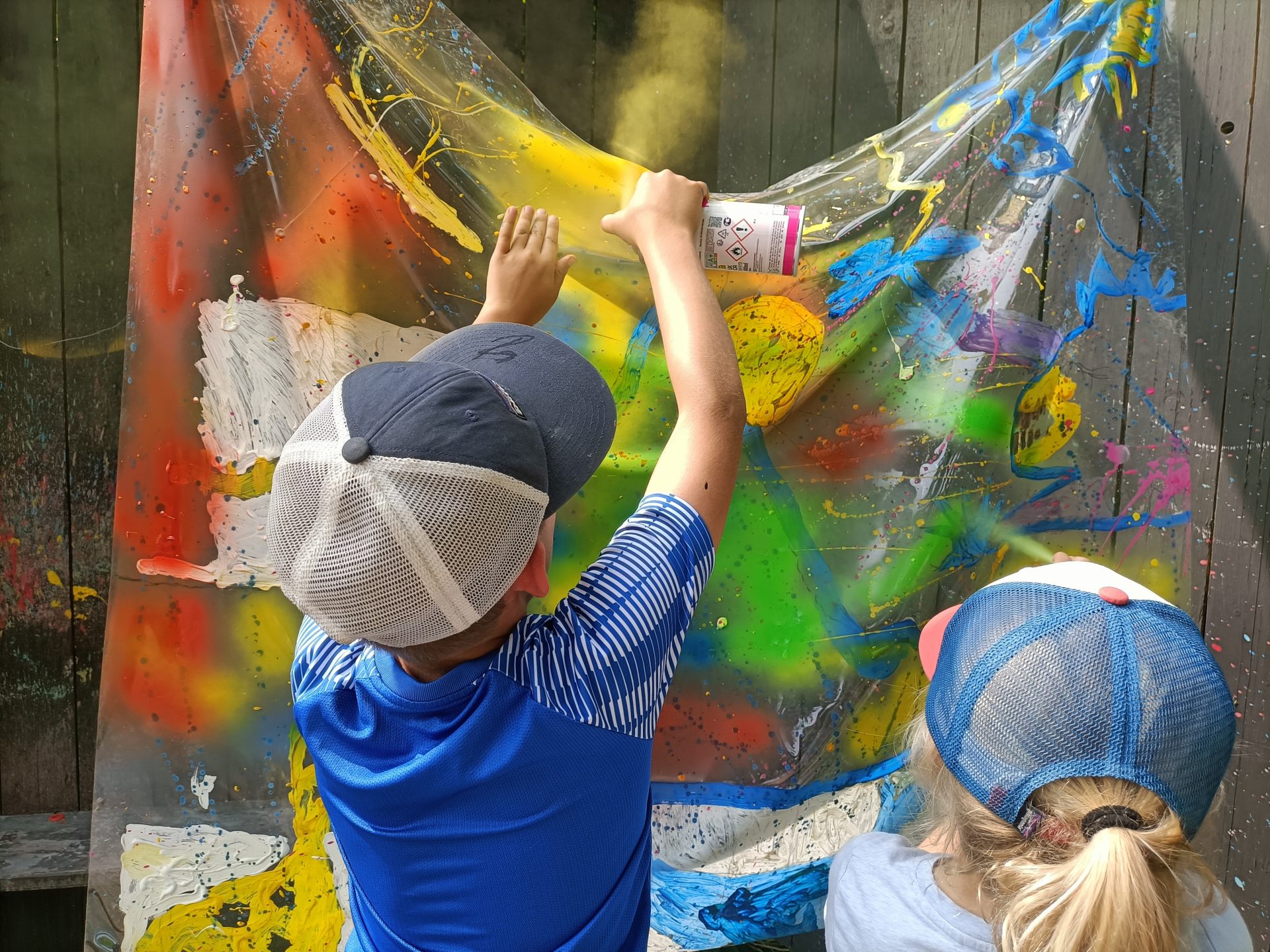 Zwei Kinder mit Baseballmützen sprühen leuchtend gelbe Farbe auf eine durchsichtige Plastikfolie, die mit bunten Spritzern bedeckt ist, und schaffen so ein Kunstprojekt im Freien vor einem Holzzaun.