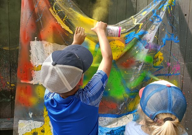 Zwei Kinder mit Baseballmützen sprühen leuchtend gelbe Farbe auf eine durchsichtige Plastikfolie, die mit bunten Spritzern bedeckt ist, und schaffen so ein Kunstprojekt im Freien vor einem Holzzaun.