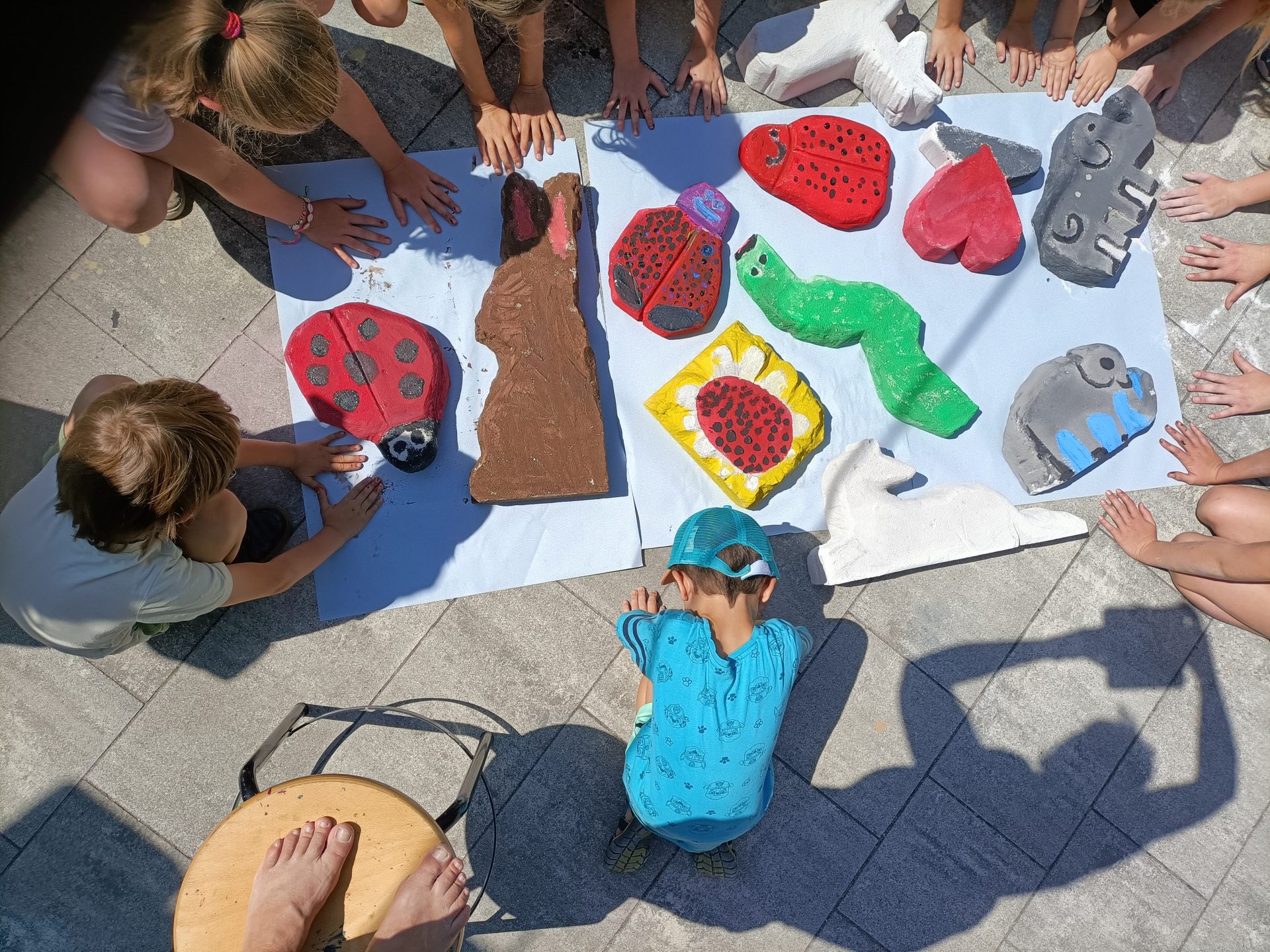 Kinder sitzen im Kreis auf einer sonnigen Terrasse und bewundern bunte, tierförmige Kunstwerke aus Ton oder Pappmaché, darunter Marienkäfer, eine Raupe und ein Elefant, die auf einem weißen Laken ausgestellt sind.