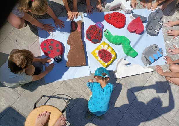 Kinder sitzen im Kreis auf einer sonnigen Terrasse und bewundern bunte, tierförmige Kunstwerke aus Ton oder Pappmaché, darunter Marienkäfer, eine Raupe und ein Elefant, die auf einem weißen Laken ausgestellt sind.