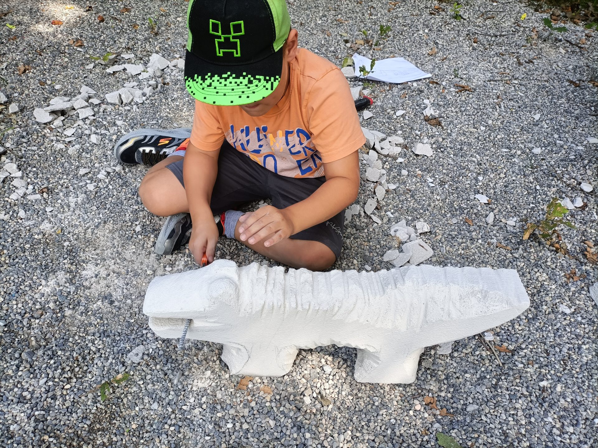 Ein Junge in orangefarbenem Hemd und grüner Mütze sitzt auf einem Kiesbett und bearbeitet mit einem Werkzeug eine weiße Steinskulptur in Form eines Tieres. Um ihn herum liegen zerbrochene Steinstücke verstreut.