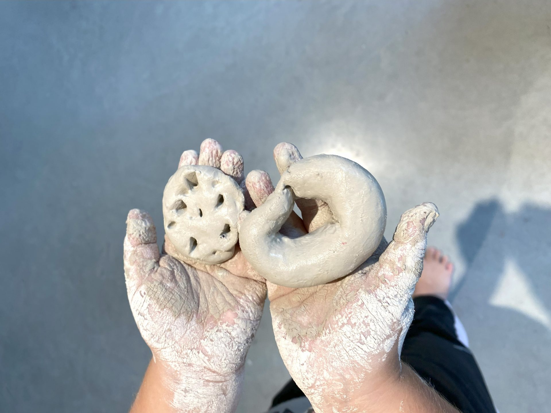 Eine Person mit lehmbedeckten Händen hält zwei kleine Tonfiguren, von denen eine die Form einer runden Scheibe mit Löchern hat und die andere einer gewundenen Form ähnelt. Der Hintergrund ist ein glatter grauer Boden.