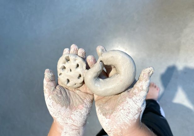 Eine Person mit lehmbedeckten Händen hält zwei kleine Tonfiguren, von denen eine die Form einer runden Scheibe mit Löchern hat und die andere einer gewundenen Form ähnelt. Der Hintergrund ist ein glatter grauer Boden.