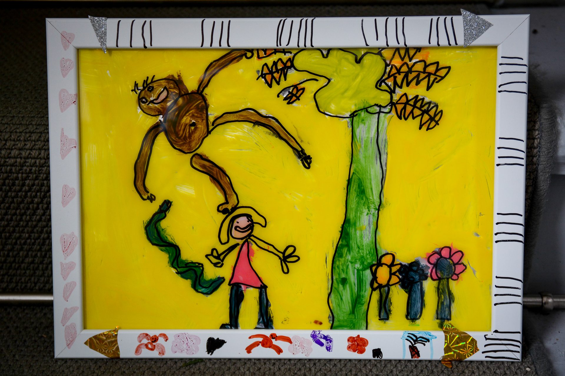 Eine farbenfrohe Kinderzeichnung in einem weiß verzierten Rahmen zeigt eine lächelnde Person unter einem hohen grünen Baum, mit einem braunen Affen, der an einem Ast hängt, und Blumen auf einem leuchtend gelben Hintergrund.