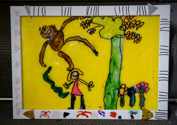 Eine farbenfrohe Kinderzeichnung in einem weiß verzierten Rahmen zeigt eine lächelnde Person unter einem hohen grünen Baum, mit einem braunen Affen, der an einem Ast hängt, und Blumen auf einem leuchtend gelben Hintergrund.