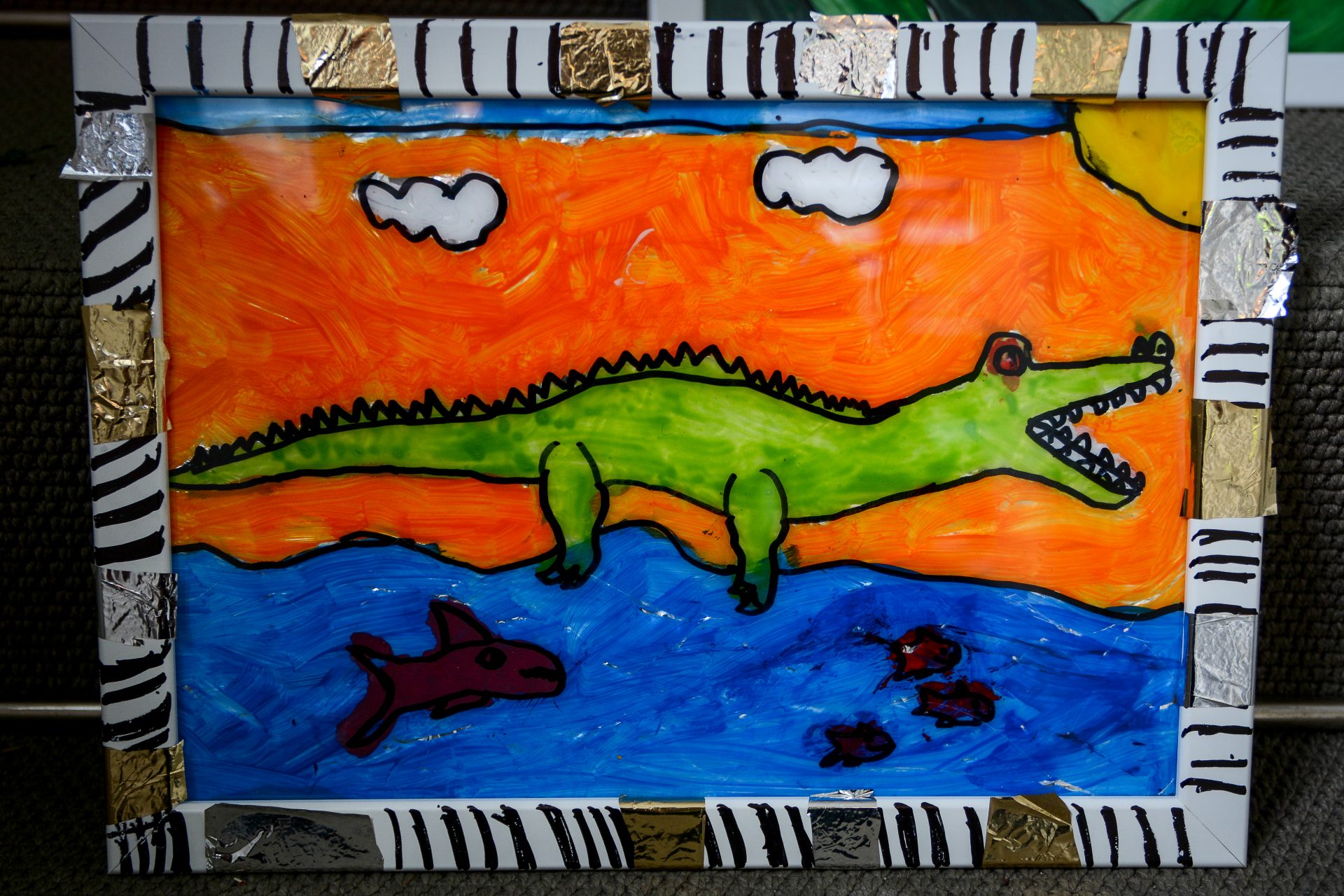 Ein farbenfrohes Kinderbild mit einem grünen Krokodil mit offenem Maul, das zwischen blauem Wasser und orangefarbenem Himmel steht, mit zwei weißen Wolken, einem lila Fisch und einem verzierten schwarz-weißen Rahmen.