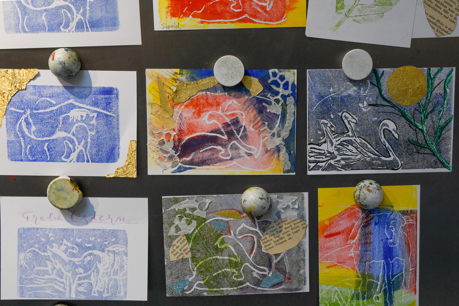 Eine Sammlung farbenfroher, handgefertigter Drucke mit Tiermotiven, darunter Pferde und Schwäne, sind mit runden, marmorartigen Magneten an eine Tafel geheftet. Einige Karten haben goldene Akzente und Collage-Elemente.