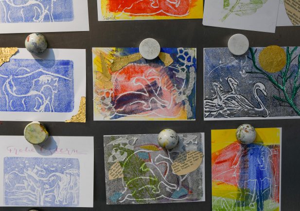Eine Sammlung farbenfroher, handgefertigter Drucke mit Tiermotiven, darunter Pferde und Schwäne, sind mit runden, marmorartigen Magneten an eine Tafel geheftet. Einige Karten haben goldene Akzente und Collage-Elemente.