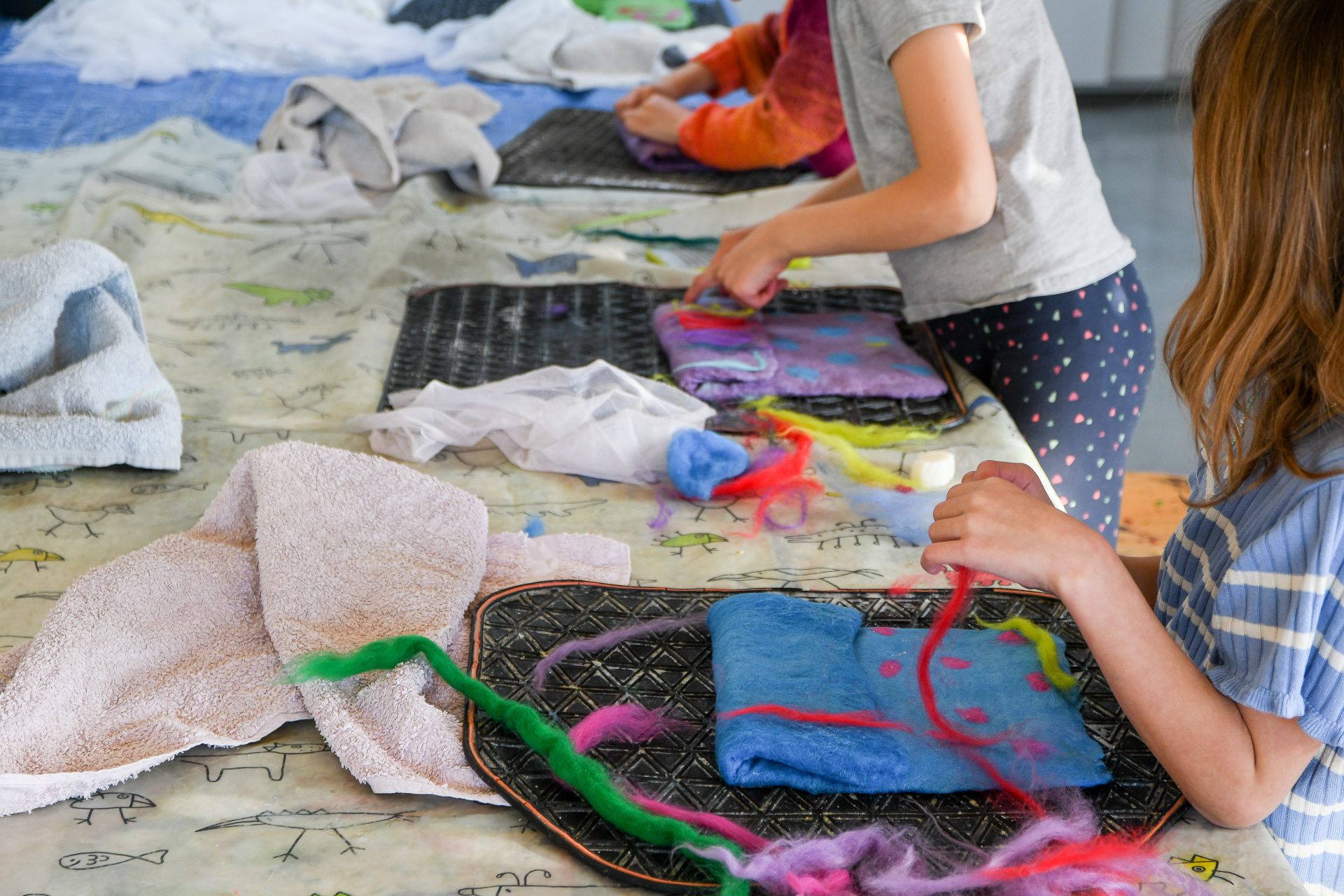 Die Kinder arbeiten an einem Bastelprojekt, bei dem sie bunte Filze und Fasern auf Tabletts auf einem Tisch anordnen, der mit einem gemusterten Tuch und einigen Handtüchern bedeckt ist. Nur ihre Arme und Hände sind sichtbar, während sie sich mit der Arbeit beschäftigen.
