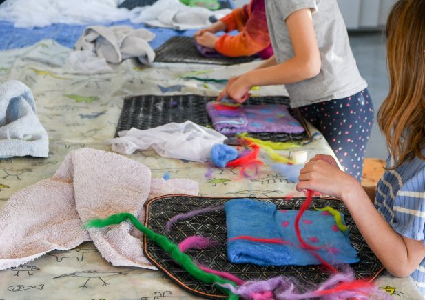 Die Kinder arbeiten an einem Bastelprojekt, bei dem sie bunte Filze und Fasern auf Tabletts auf einem Tisch anordnen, der mit einem gemusterten Tuch und einigen Handtüchern bedeckt ist. Nur ihre Arme und Hände sind sichtbar, während sie sich mit der Arbeit beschäftigen.