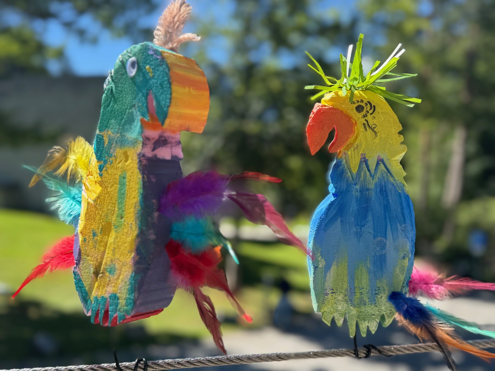 Zwei bunte, handgefertigte Papagei-Dekorationen aus bemaltem Papier und Federn sitzen auf einem Draht im Freien, mit einem verschwommenen grünen Hintergrund und Bäumen in der Ferne.