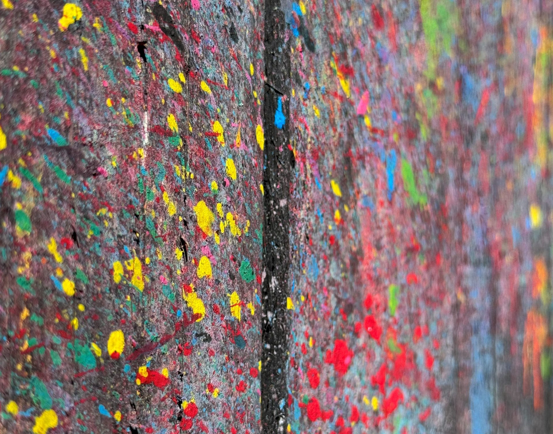 Nahaufnahme einer Betonwand, die mit leuchtenden Farben, darunter Gelb, Rot, Grün und Blau, bespritzt ist, was ihr ein lebendiges, abstraktes Aussehen verleiht. Eine vertikale Linie unterteilt die Wand in zwei Abschnitte.